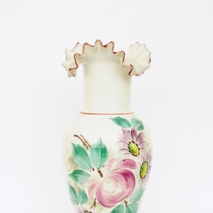 Puede incluir: Un jarrón de cerámica blanca con un borde ondulado y un diseño floral pintado. El jarrón presenta flores rosas y moradas con hojas verdes.