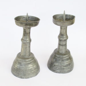 May include: Two antique pewter candle holders with a simple design. Each holder has a round base with a fluted design, a cylindrical stem, and a round cup for the candle.