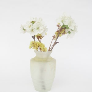 Puede incluir: Un jarrón de cerámica blanco con un acabado mate contiene un ramo de flores blancas y amarillas. Las flores están dispuestas en un estilo simple y elegante.