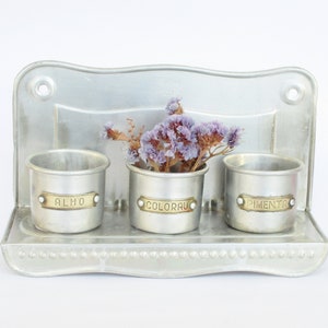 May include: A silver metal wall-mounted spice rack with three small containers labeled "ALHO", "COLORAU", and "PIMENTA".