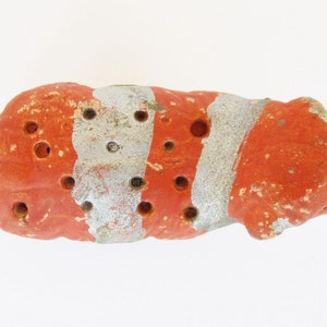 May include: A small, red and white striped ceramic object with multiple holes. The object is irregularly shaped and has a rough texture.