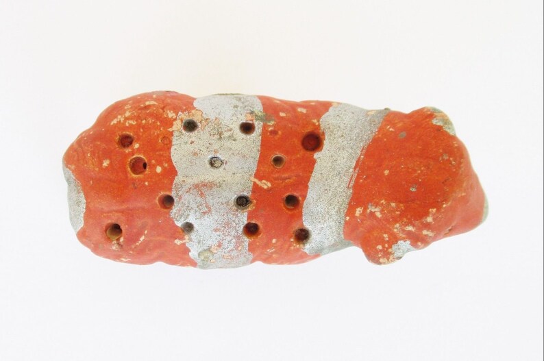 May include: A small, red and white striped ceramic object with multiple holes. The object is irregularly shaped and has a rough texture.