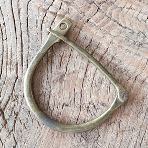 Puede incluir: Un anillo de lat&oacute;n en forma de D con un borde redondeado. El anillo es liso y tiene un acabado ligeramente desgastado.