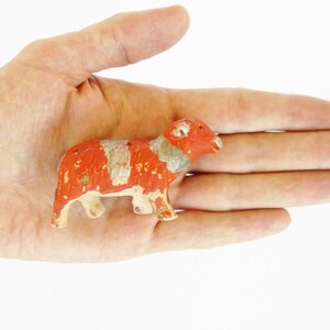 May include: A small, red and white painted ceramic figurine of a ram with a curled horn. The figurine is standing on its four legs and has a detailed face.