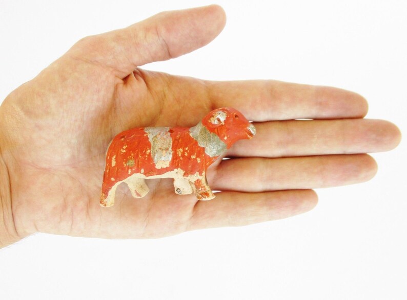 May include: A small, red and white painted ceramic figurine of a ram with a curled horn. The figurine is standing on its four legs and has a detailed face.