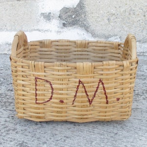 May include: A woven wicker basket with two handles, featuring the letters "D.M." written in red paint on the front.