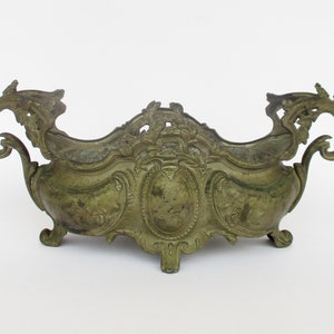 May include: An antique brass planter with ornate scrollwork and a decorative design. The planter has two handles and sits on three feet.