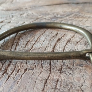 Puede incluir: Un brazalete de lat&oacute;n con un dise&ntilde;o simple y curvo y una peque&ntilde;a abertura redonda en un lado.