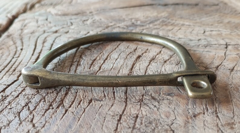Puede incluir: Un brazalete de lat&oacute;n con un dise&ntilde;o simple y curvo y una peque&ntilde;a abertura redonda en un lado.