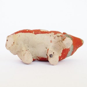 May include: A small ceramic pig figurine with red, white, and gray paint. The pig is lying on its side with its legs visible.