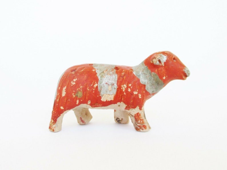 May include: A small, red and white ceramic sheep figurine. The sheep is standing on all four legs and has a slightly tilted head. The figurine is painted with a red base and white accents.