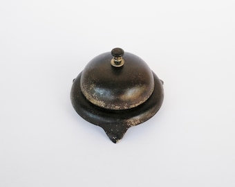 May include: A dark bronze bell with a small, rounded top and a wider base. The bell has a tarnished, aged appearance, suggesting it is an antique. The base has a decorative, three-point design. A classic item.