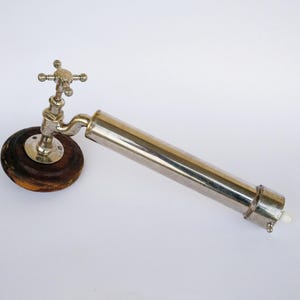 Peut inclure: Une pompe à eau de style antique en métal argenté avec une base en bois. La pompe a un long fût cylindrique et un robinet avec une poignée en forme de croix.