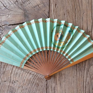 May include: A green paper fan with a gold and floral border. The fan features a painted scene of two figures in a circle.