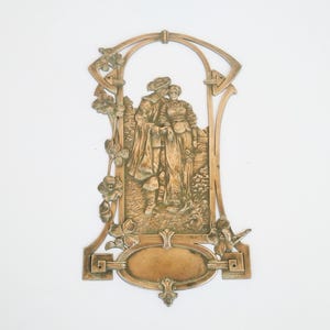 May include: A bronze-toned wall plaque featuring a relief of a couple in period clothing, framed by an arched design with floral and geometric elements. The plaque includes an oval space for personalization. The overall design suggests a vintage or antique aesthetic.