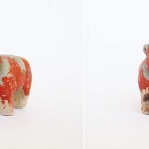 May include: A small, hand-painted ceramic figurine of a red and white cow. The cow is standing on all four legs and has a simple, cartoonish design.