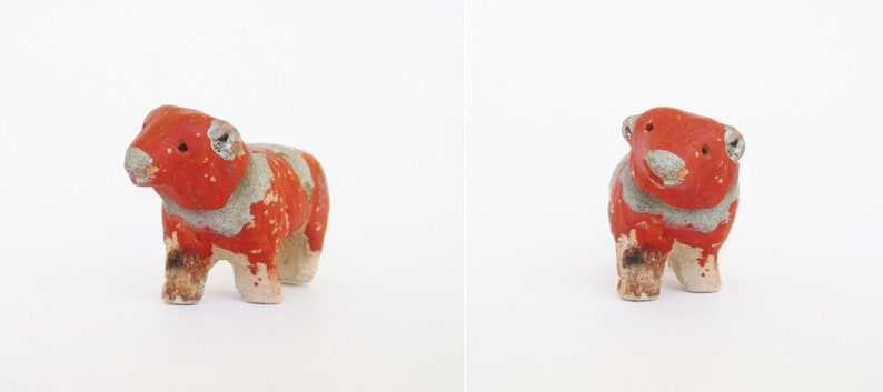 May include: A small, hand-painted ceramic figurine of a red and white cow. The cow is standing on all four legs and has a simple, cartoonish design.