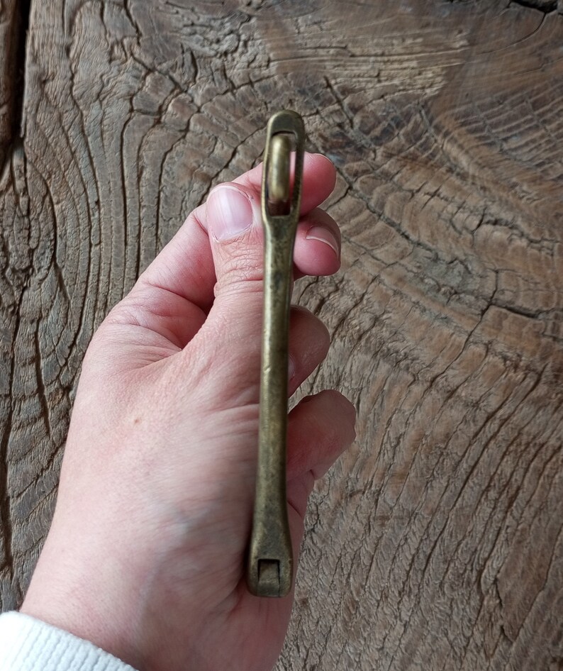 Puede incluir: Un tirador de caj&oacute;n de lat&oacute;n con un dise&ntilde;o simple y recto. El tirador tiene una parte superior redondeada y una base rectangular.