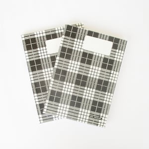 May include: Two black and white plaid notebooks with a blank label on the front. The notebooks are stacked on top of each other.