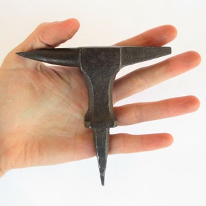 Puede incluir: Un pequeño yunque antiguo de hierro con un cuerno puntiagudo y una parte superior plana. El yunque se sostiene en una mano para mostrar su tamaño.