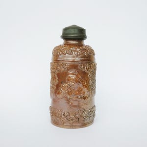 May include: A brown salt-glazed stoneware bottle featuring a dark green metal cap. The cylindrical body displays embossed relief carvings of a person drinking from a mug, surrounded by intricate grapevines and foliage patterns on the top and bottom.
