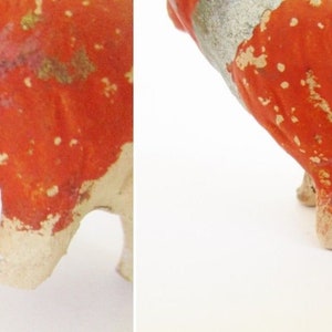 May include: A ceramic pig figurine with red and white paint. The pig is standing on all four legs and has a chipped paint finish.