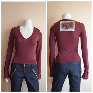 May include: A maroon long-sleeve shirt with a v-neck and gold buttons on the cuffs. The shirt has a small embroidered patch on the back that says "LIVE". The shirt is being modeled on a mannequin wearing blue jeans.