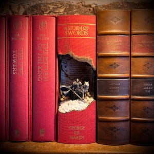 May include: A display of hardback books with red and brown covers. One red book features a carved-out scene of a figure with a wolf. The books are titled "The Two Towers", "The Return of the King", and "A Storm of Swords".