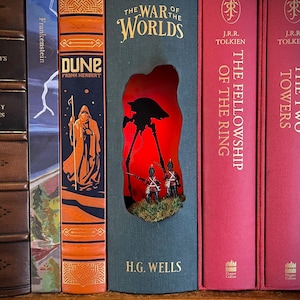 May include: A book nook insert featuring a dark green spine titled The War of the Worlds by H.G. Wells. The center is hollowed out to reveal a red-lit diorama containing a black tripod machine silhouette and two miniature soldiers on grass.