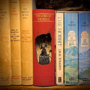 Peut inclure: Une collection de livres de J.R.R. Tolkien, dont "Le Seigneur des Anneaux" et "Le Hobbit". Les livres sont de couleurs variées, avec le volume rouge "Le Seigneur des Anneaux" présentant une découpe révélant une illustration. Les livres sont disposés sur une étagère.