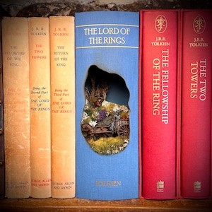 May include: A collection of J.R.R. Tolkien's "The Lord of the Rings" books. The spines are in various colors, including beige, red, and blue. The center book has a cutout revealing a miniature diorama of a landscape with a horse-drawn cart.