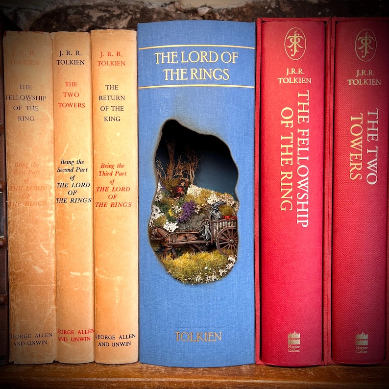 May include: A collection of J.R.R. Tolkien's "The Lord of the Rings" books. The spines are in various colors, including beige, red, and blue. The center book has a cutout revealing a miniature diorama of a landscape with a horse-drawn cart.