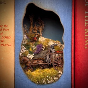 May include: A diorama scene from a book, featuring a horse-drawn cart with a wizard figure, a hobbit, and a landscape of flowers and trees. The scene is set within a cutout on a blue background, with the spine of a book visible on either side.