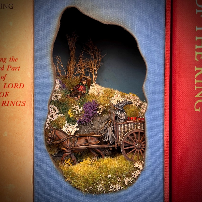 May include: A diorama scene from a book, featuring a horse-drawn cart with a wizard figure, a hobbit, and a landscape of flowers and trees. The scene is set within a cutout on a blue background, with the spine of a book visible on either side.