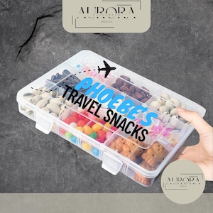 May include: A clear plastic snack box, divided into compartments, containing an assortment of treats such as nuts, sweets, and biscuits. The box is labelled "PHOEBE'S TRAVEL SNACKS" with an aeroplane illustration.
