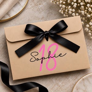 May include: A rectangular tan gift box featuring a black satin ribbon tied in a bow at the top centre. The front of the box displays the name Sophie in black cursive script, layered over a large, bright pink number 18 printed on the surface.