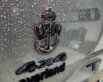 May include: A gold-colored USN anchor emblem with a chain, mounted on a gray vehicle. Below the anchor is a black 4x4 Overland emblem. The vehicle is covered in raindrops.