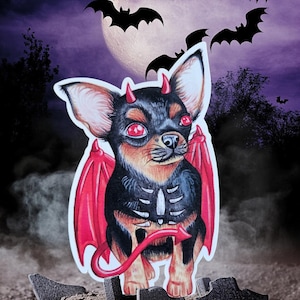 May include: A black and brown chihuahua dressed as a devil with red wings and a tail. The dog is standing on a pile of rocks in front of a full moon and bats.
