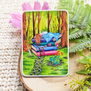 May include: A rectangular sticker with a fantasy illustration of a person asleep on a pile of books in a forest. A small dragon is resting nearby. A sword and a small ladder are also present. The sticker is on a wooden surface.