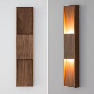 Op de afbeelding: Een moderne wandlamp met een rechthoekig houten frame en twee lichtpanelen. De lamp is in de tweede afbeelding verlicht, wat de warme gloed van het licht laat zien.