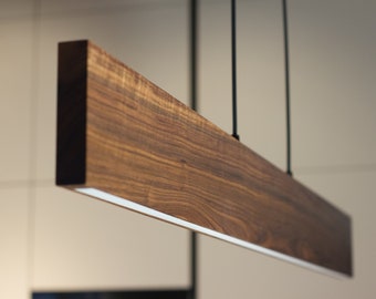 May include: A modern wooden pendant light fixture with a sleek, rectangular design. The light is suspended from the ceiling by black cords and features a warm wood finish.