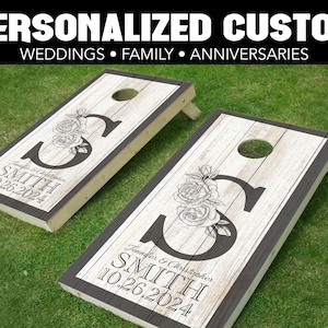 May include: Two personalized wooden cornhole boards with a black border. The boards feature a whitewashed wood background with a black initial "S" and floral design. The text "Jennifer & Christopher Smith 10.26.2024" is printed on each board.