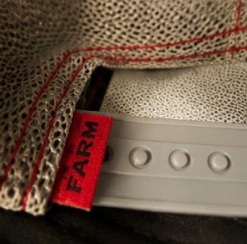 May include: Close-up of a gray baseball cap with a mesh top and a red tag that reads "FARM". The cap has a gray brim with adjustable snaps.