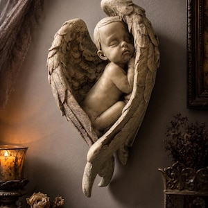 May include: A beige-colored wall sculpture of a sleeping baby nestled within angel wings. The sculpture has a textured finish and is mounted on a gray wall. The wings are detailed with feather patterns, creating a serene and artistic piece.