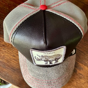 May include: A gray and brown baseball cap with a leather front panel and a red button on top. The cap features a patch with a rhino illustration and the word "TOUGH".