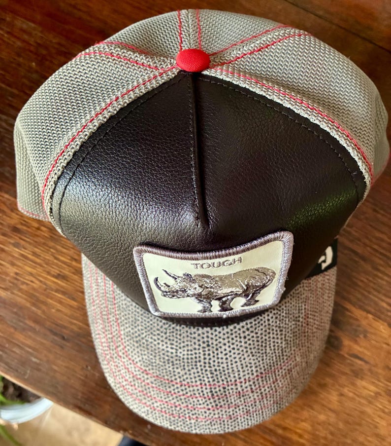 May include: A gray and brown baseball cap with a leather front panel and a red button on top. The cap features a patch with a rhino illustration and the word "TOUGH".