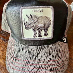 May include: A black and gray trucker hat with a rhino patch. The patch features a detailed illustration of a rhino with the word "TOUGH" above it. The hat has a black leather-like front panel and a gray dotted mesh back with red stitching.