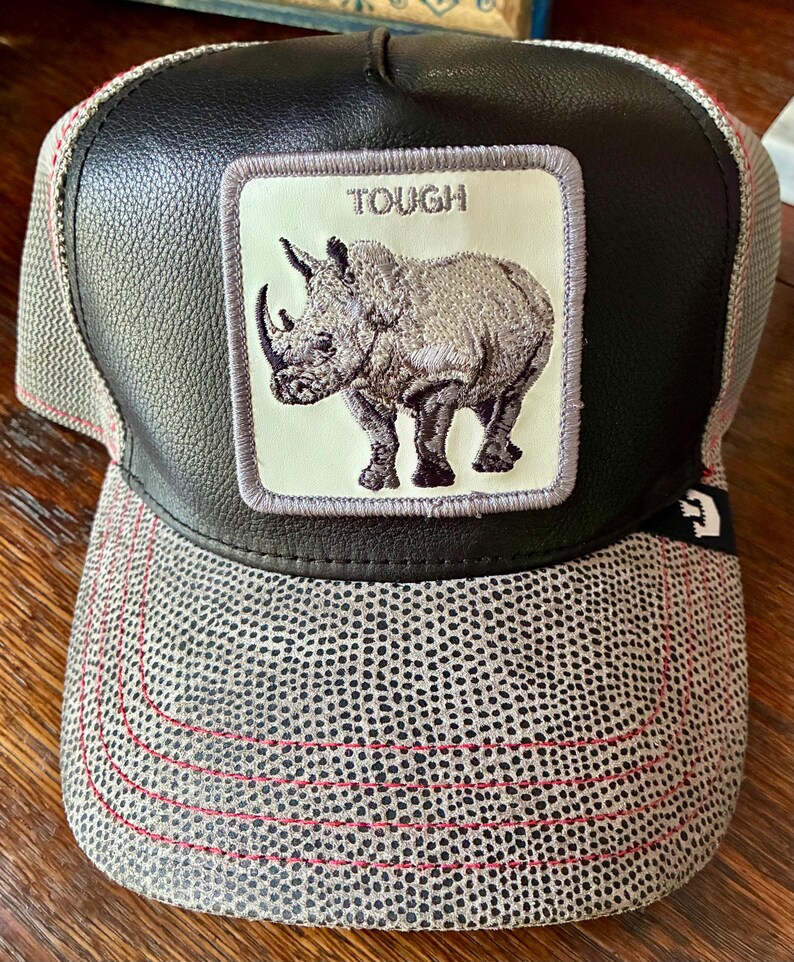 May include: A black and gray trucker hat with a rhino patch. The patch features a detailed illustration of a rhino with the word "TOUGH" above it. The hat has a black leather-like front panel and a gray dotted mesh back with red stitching.