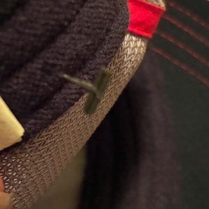 May include: Close-up of a dark purple knitted fabric with a textured, woven lining. A small, dark green plastic tag is attached to the fabric. A red fabric edge is visible.
