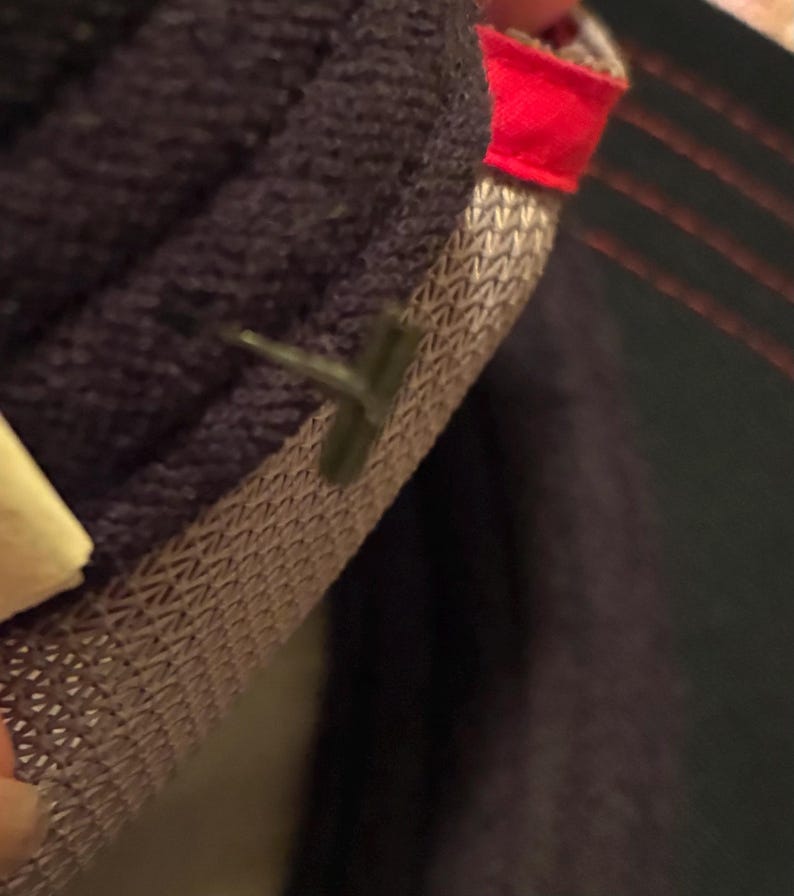 May include: Close-up of a dark purple knitted fabric with a textured, woven lining. A small, dark green plastic tag is attached to the fabric. A red fabric edge is visible.